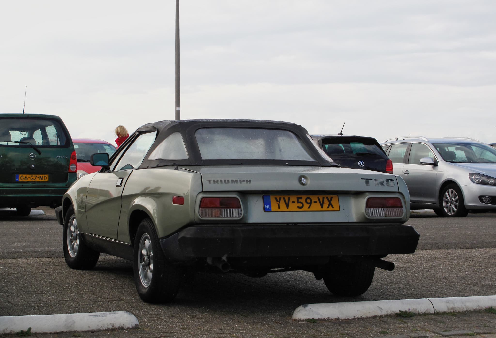 A silver-green TR8 convertible from behind in a car park, its hood up, with raised TR8 letters and a tailpipe at each side.