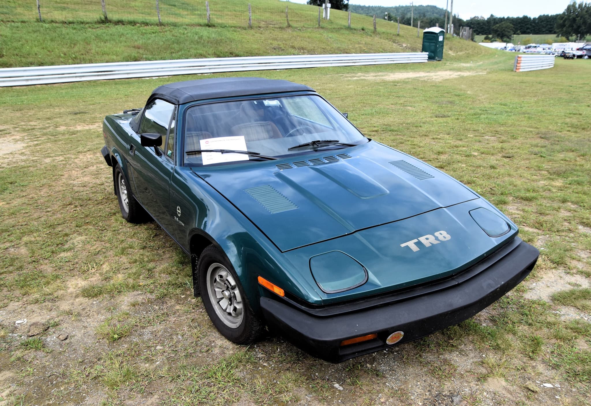 A dark green Triumph TR8 convertible with its black hood up, parked on grass beside a circuit's guard rail.